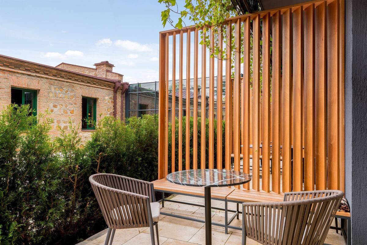 A patio with a glass-top table and two wicker chairs, surrounded by a wooden fence and greenery.