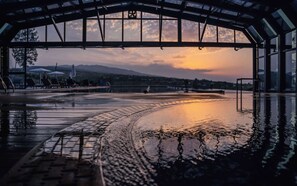 An indoor swimming pool with a glass ceiling, a view of the sunset, and a few lounge chairs.