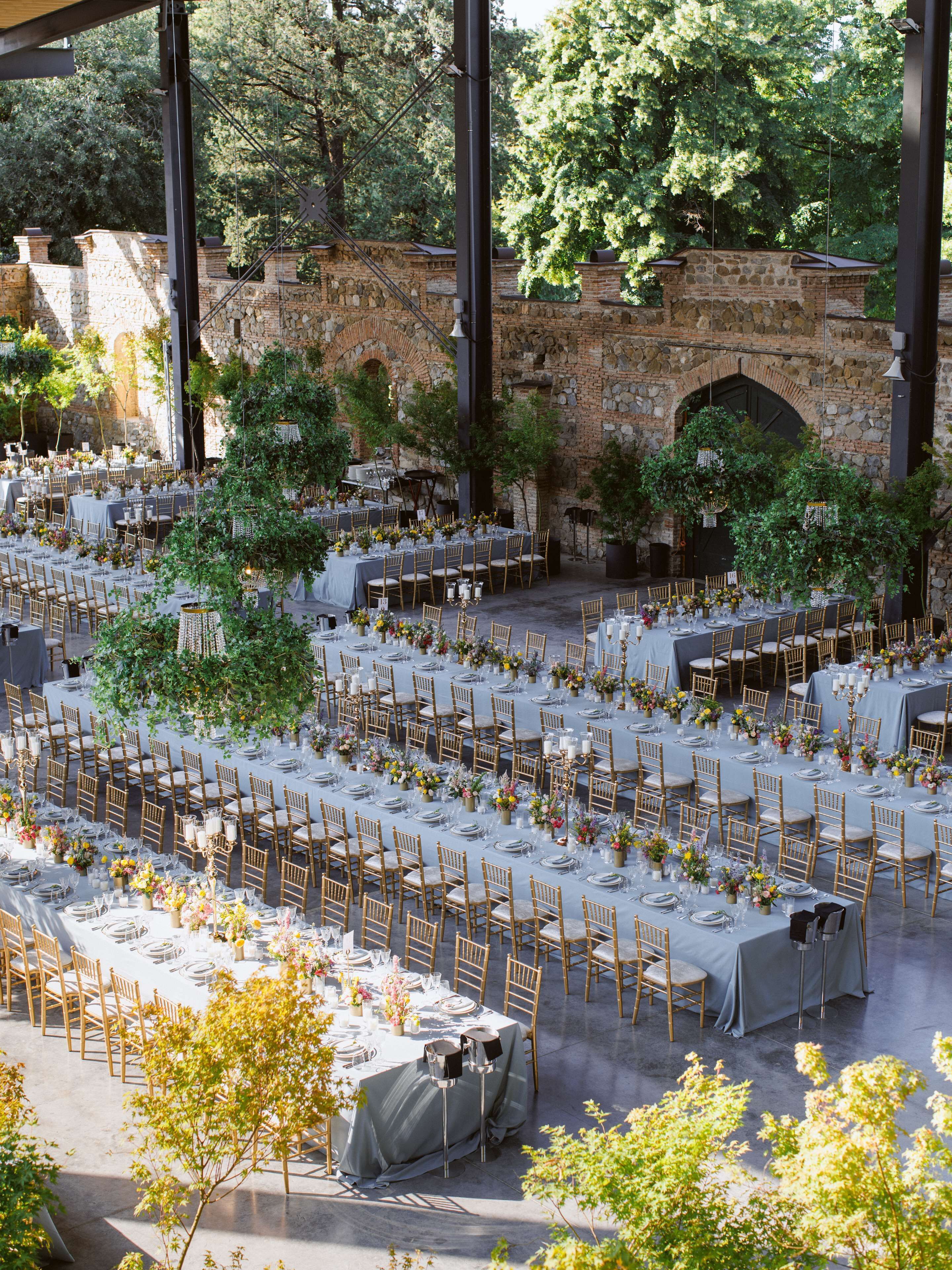 An outdoor banquet hall with multiple round tables set for dining, surrounded by stone walls and greenery.