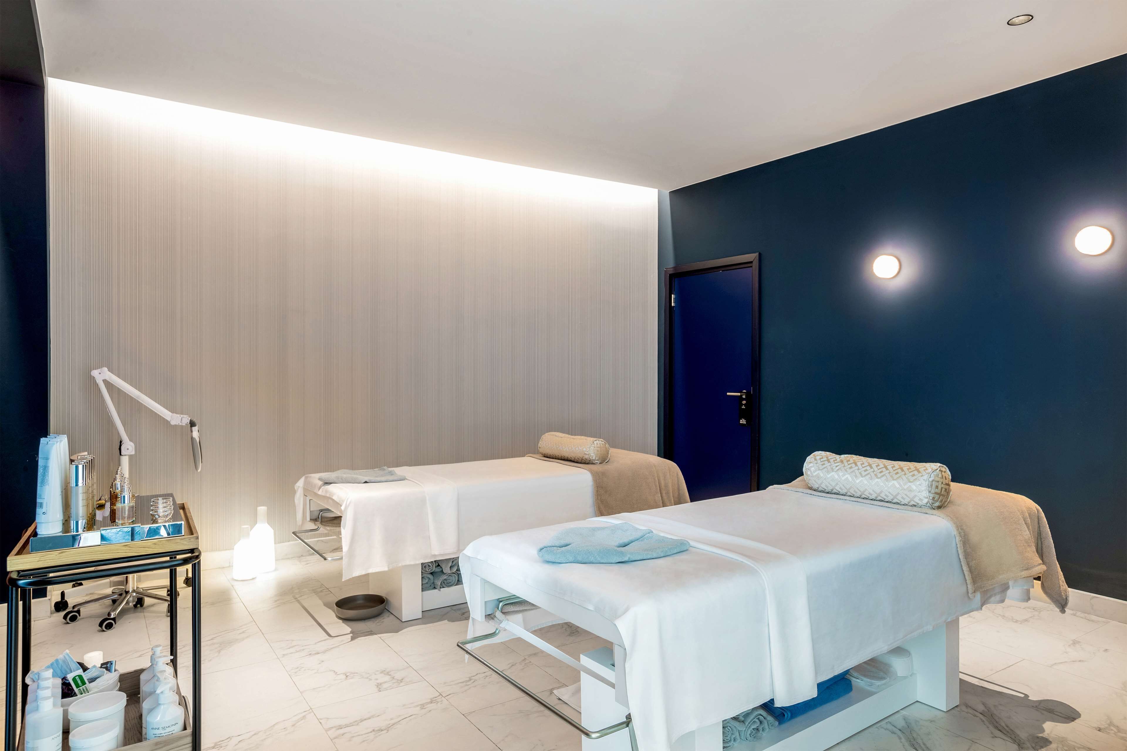A modern spa room with two treatment beds, a massage table, and a cart with supplies.
