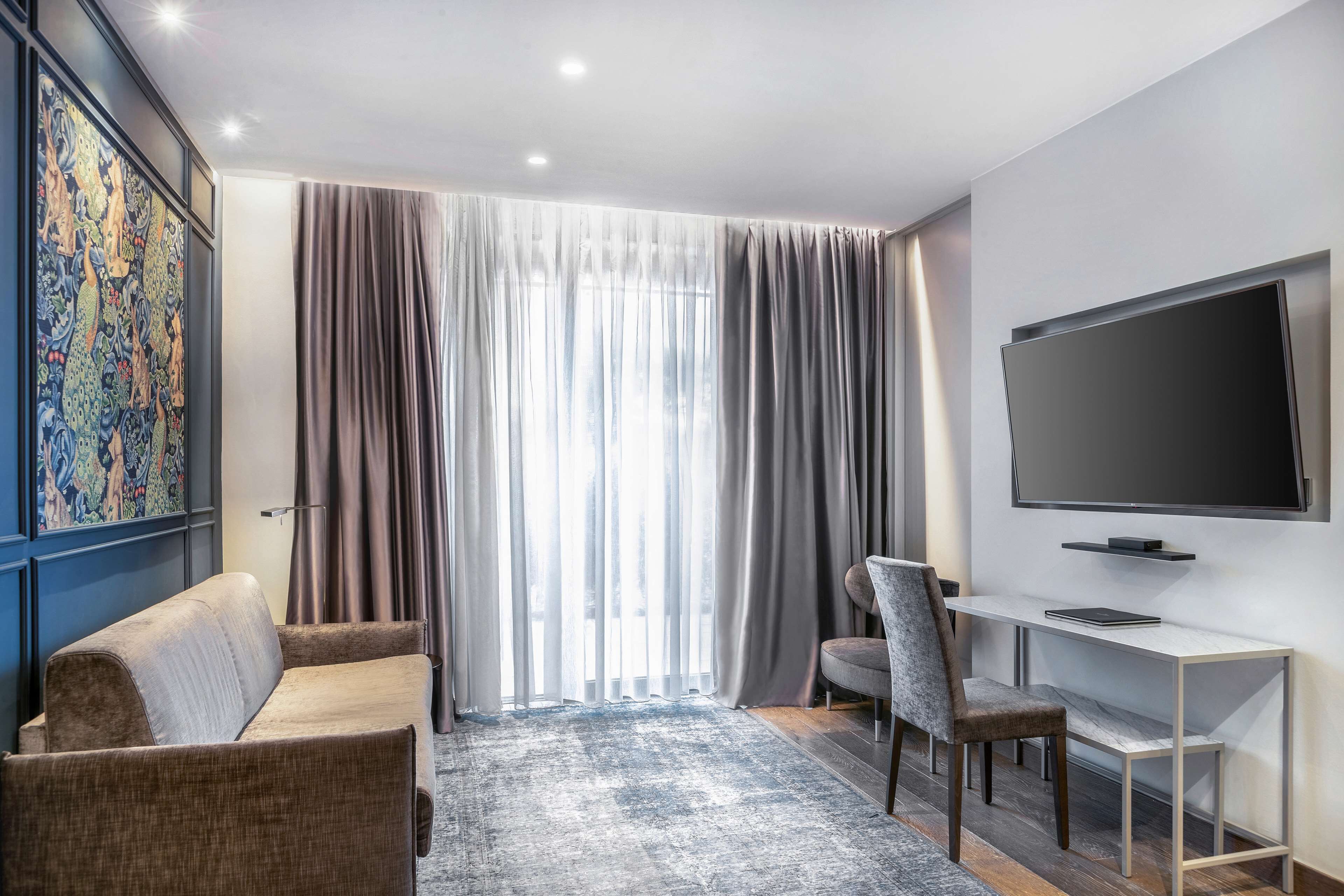 Una habitación de hotel moderna con un ventanal grande, un televisor de pantalla plana, un sofá, una silla y un escritorio.