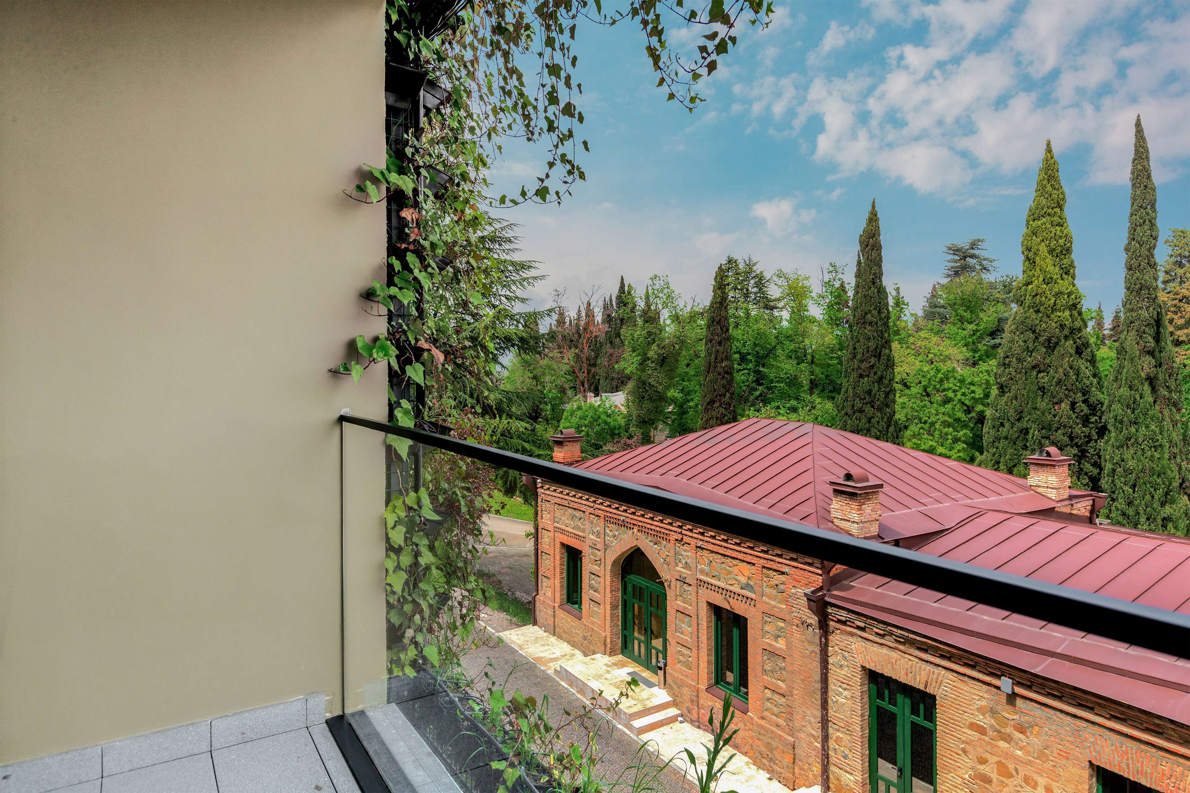 Une vue depuis un balcon donnant sur un bâtiment traditionnel en briques avec un toit rouge, entouré de verdure et de grands arbres.