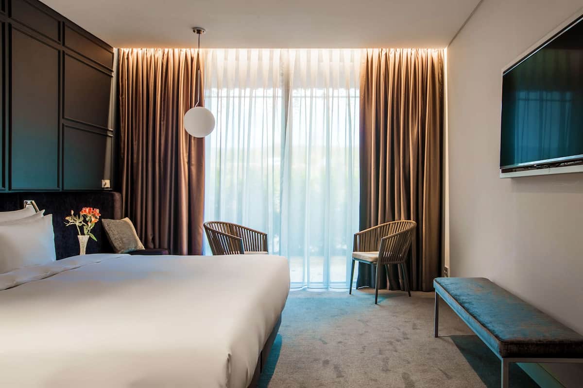 A hotel room with a large bed, two chairs, a bench, a TV, and a window with curtains.
