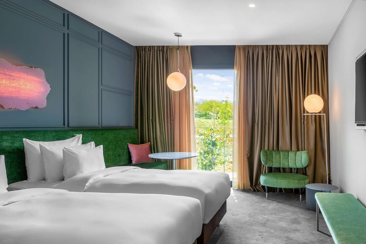 A hotel room with a large bed, a green upholstered chair, a small round table, and a view of greenery outside.