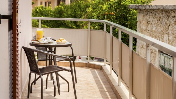 A balcony with a table and chairs, a glass of orange juice, and a view of greenery and buildings.
