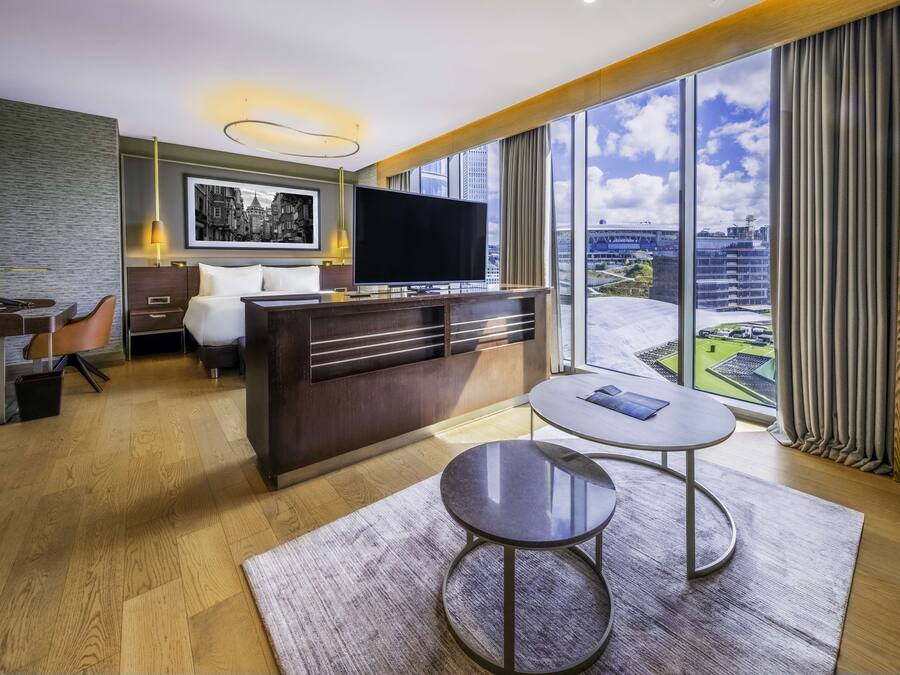 Habitación de hotel moderna con una cama grande, un escritorio, un televisor de pantalla plana y vistas de la ciudad a través de amplios ventanales.