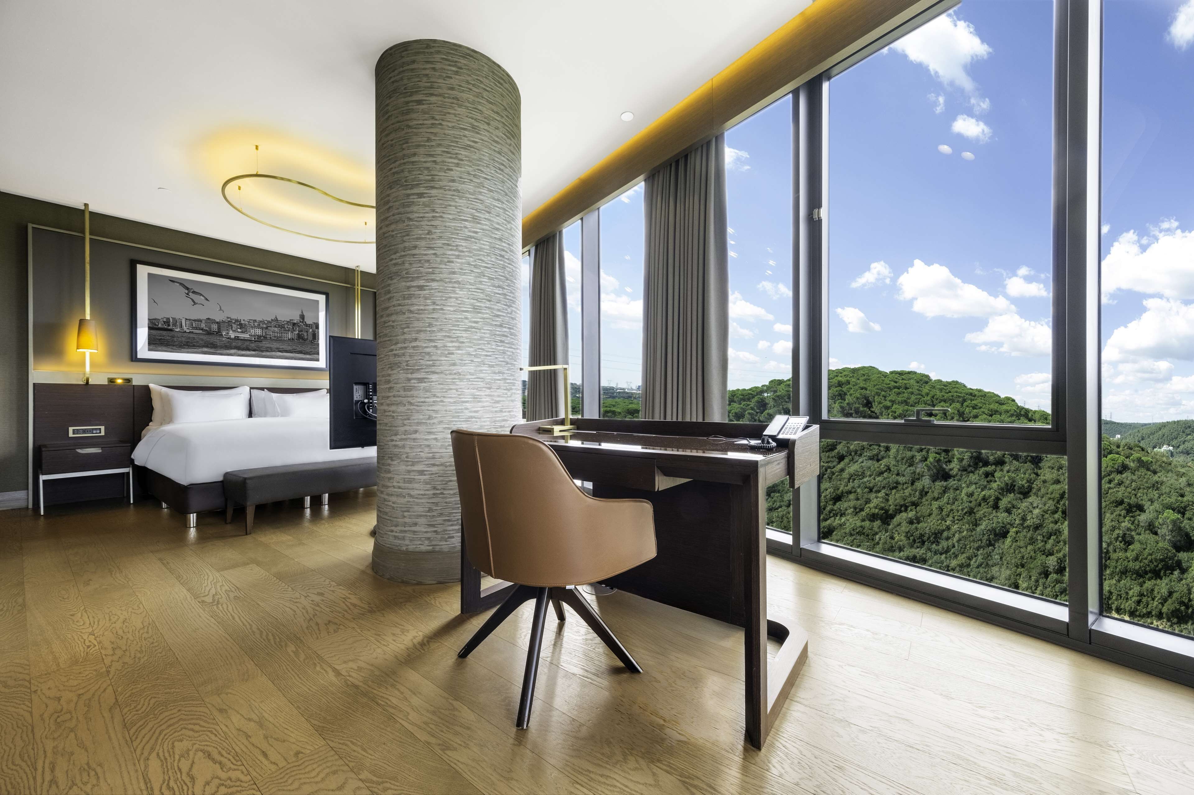 A modern hotel room with a large desk, a comfortable bed, a large window with a cityscape view, and a large pillar.