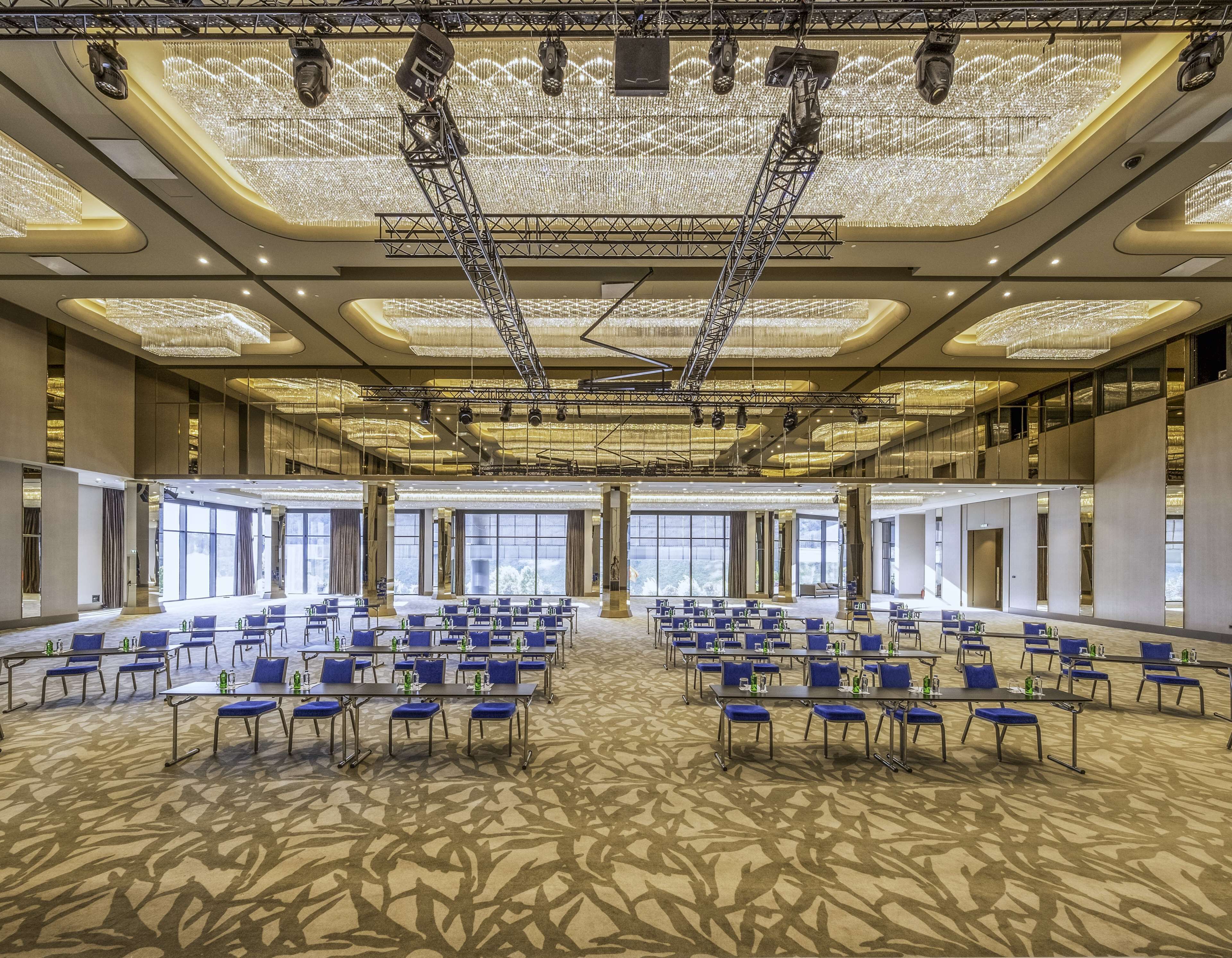 Un amplio salón de conferencias con filas de mesas y sillas, ventanas grandes y un techo alto con candelabros.