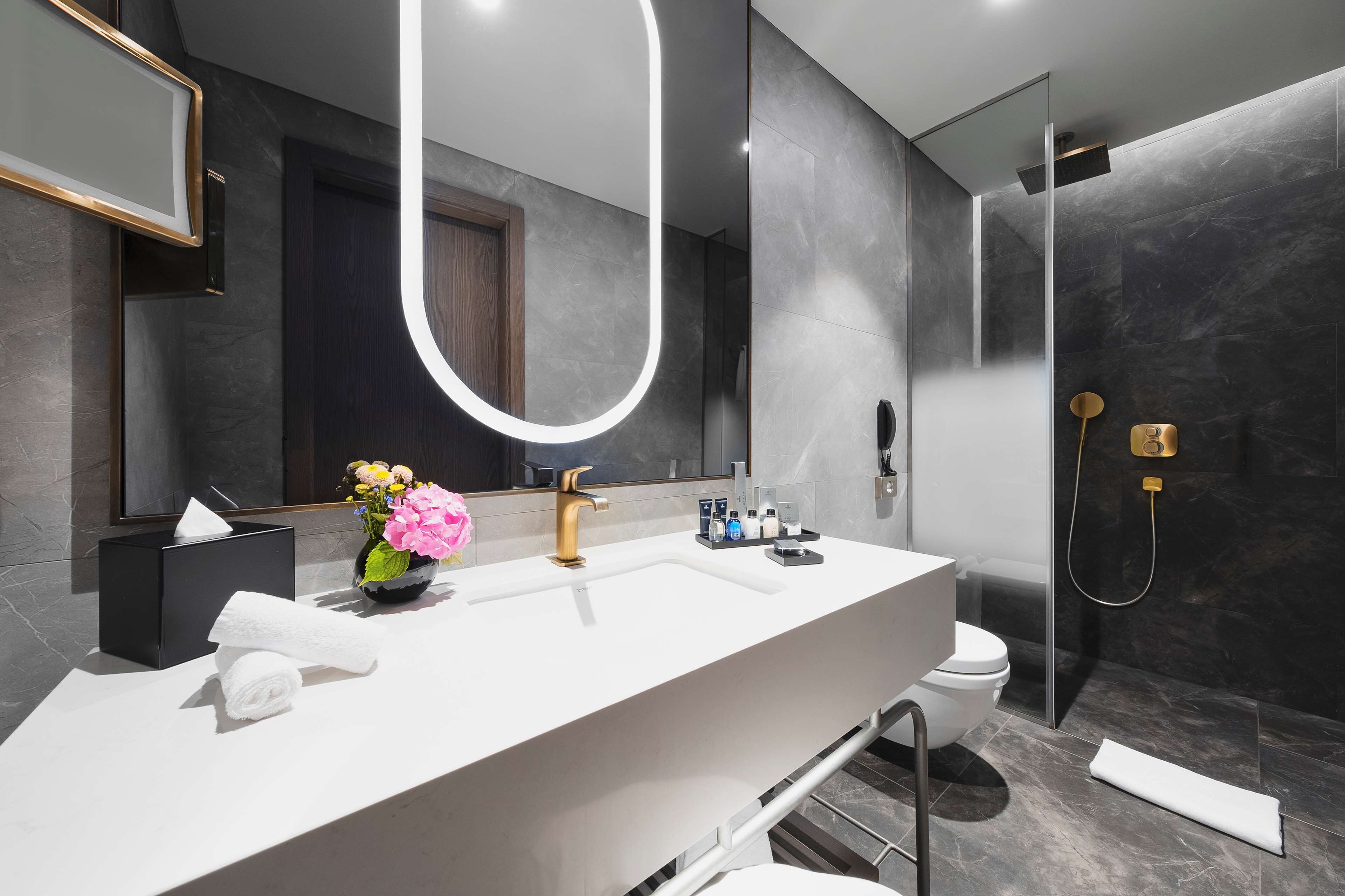 A modern bathroom with a white vanity, a large mirror, a gold faucet, and a shower area.