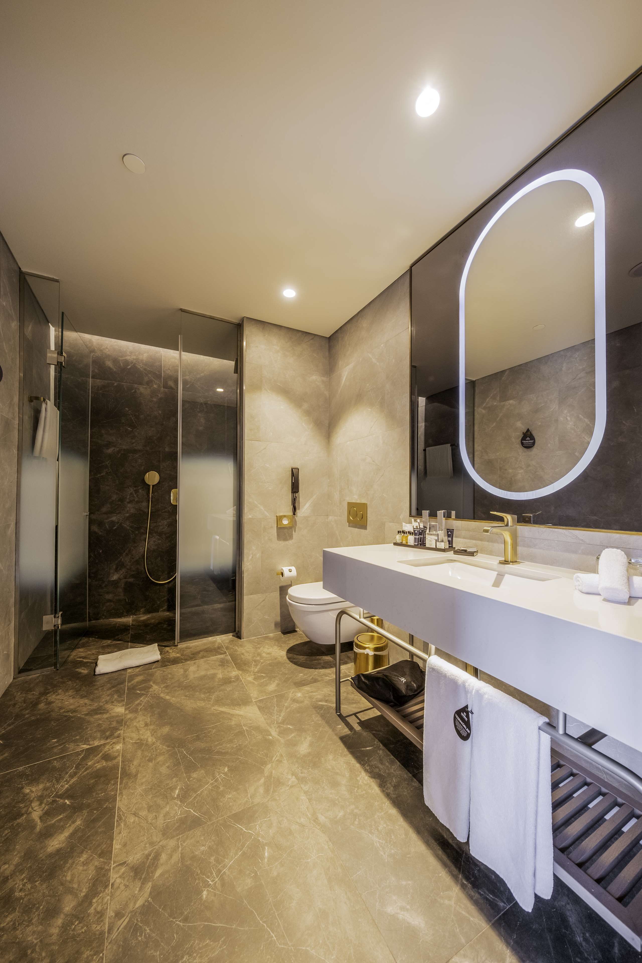 Un bagno moderno con doccia vetrata, lavandino bianco con dettagli dorati e un grande specchio con cornice illuminata.
