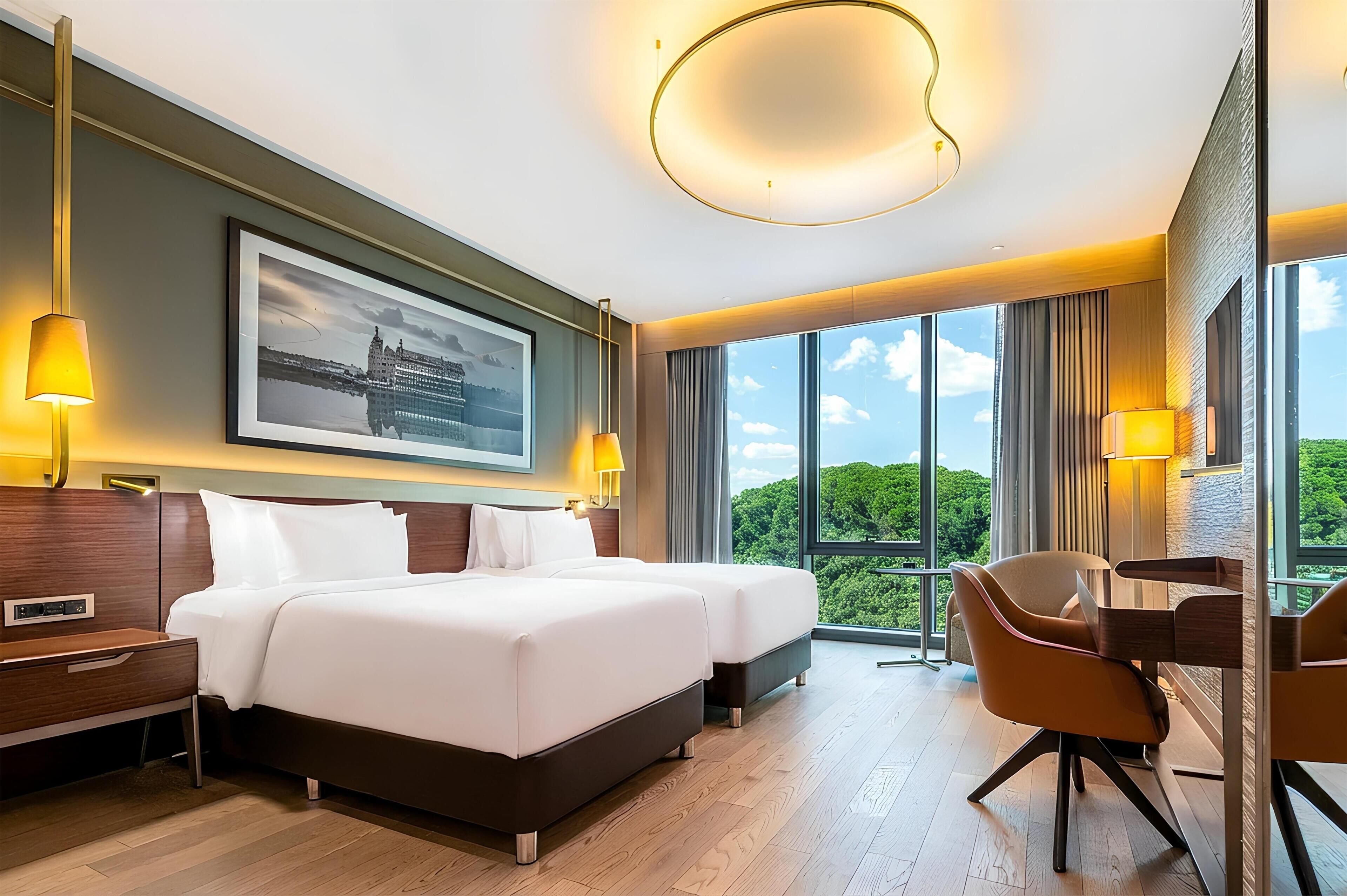 A hotel room with a large bed, two bedside tables, a desk with a chair, and a large window offering a view of greenery.