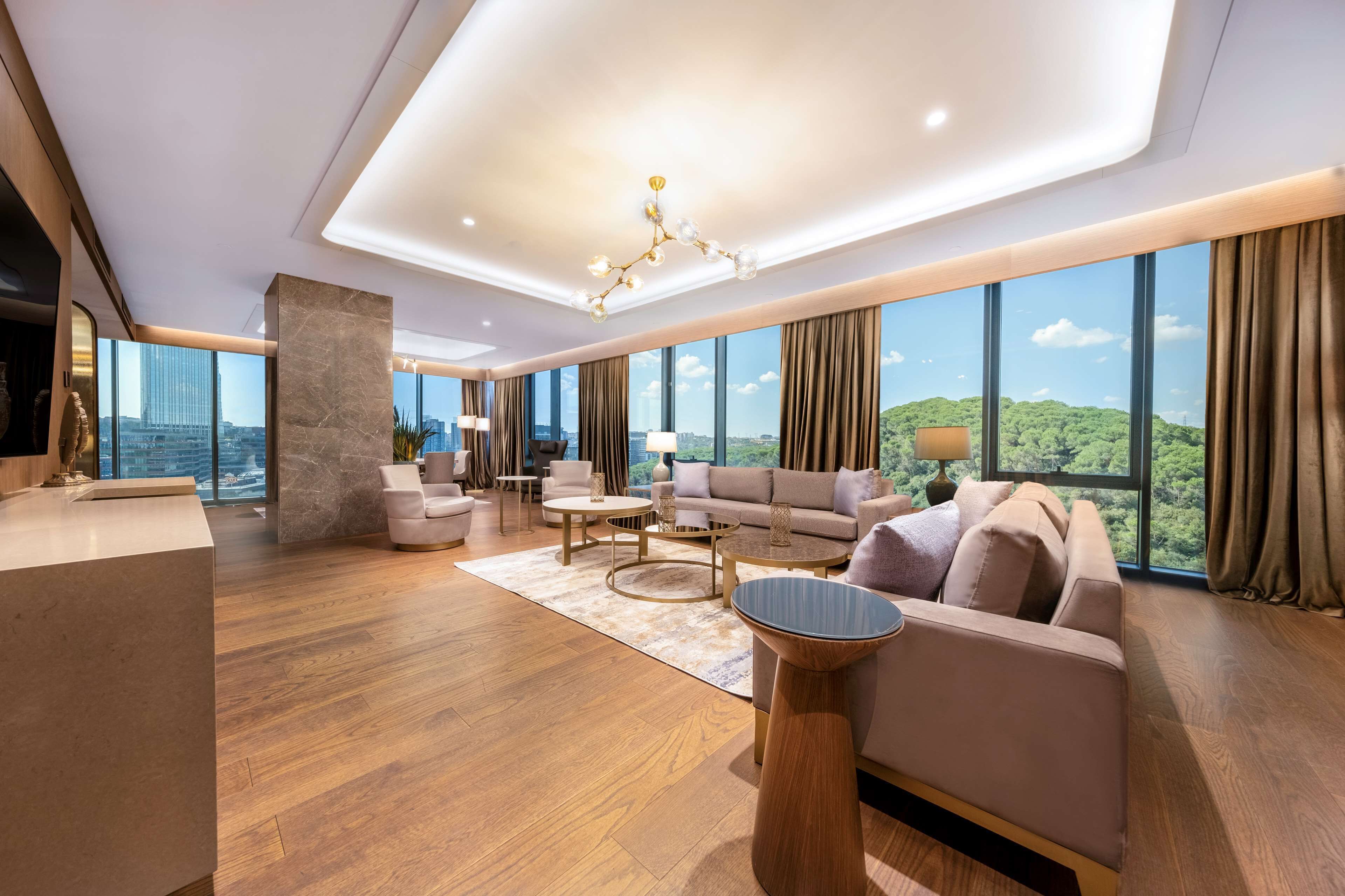 A spacious living room with a large window offering a city view, a marble pillar, and a modern chandelier.