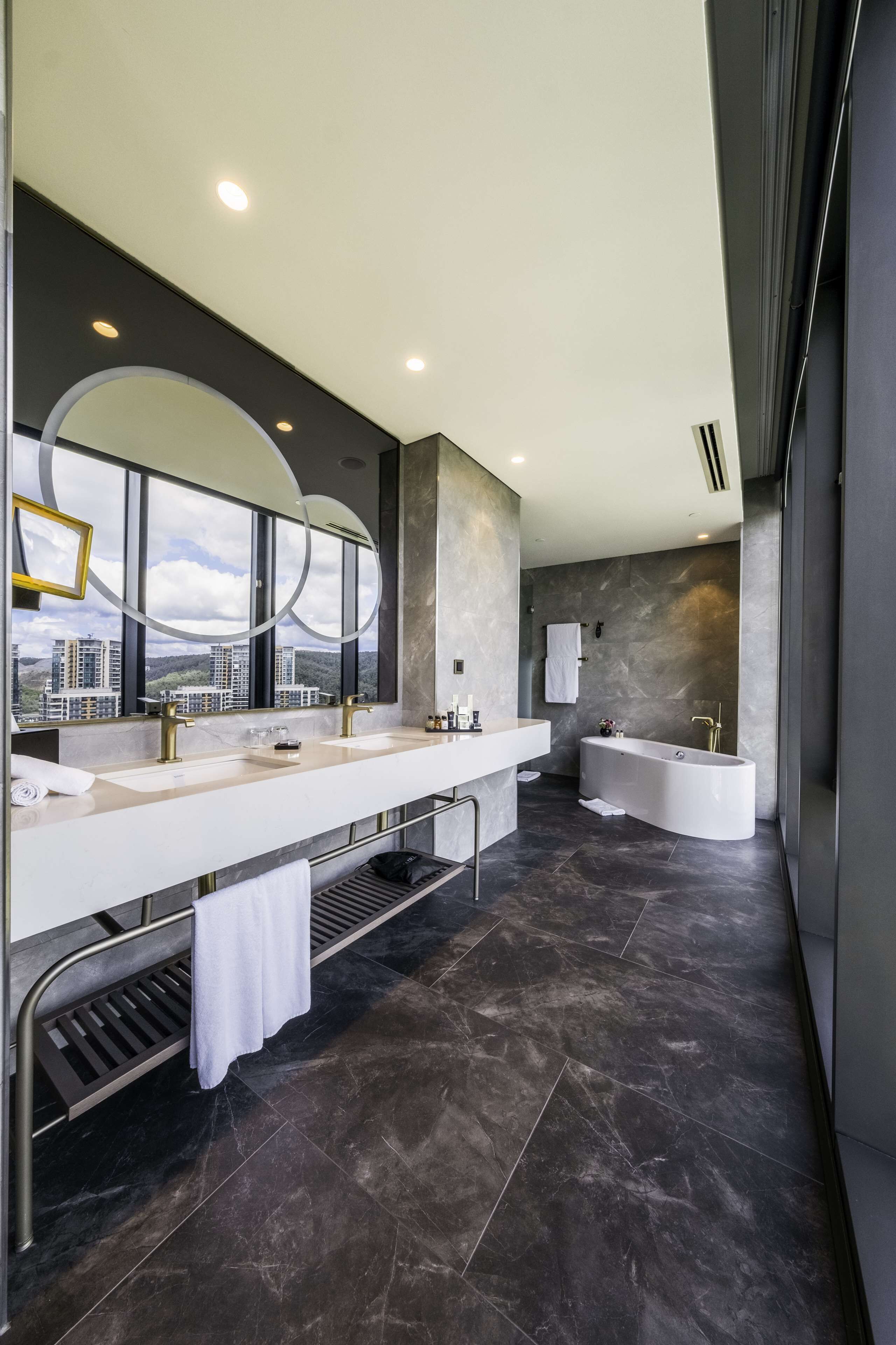 Een moderne badkamer met een ruime wastafel, een vrijstaand bad en uitzicht op de stad door grote ramen.