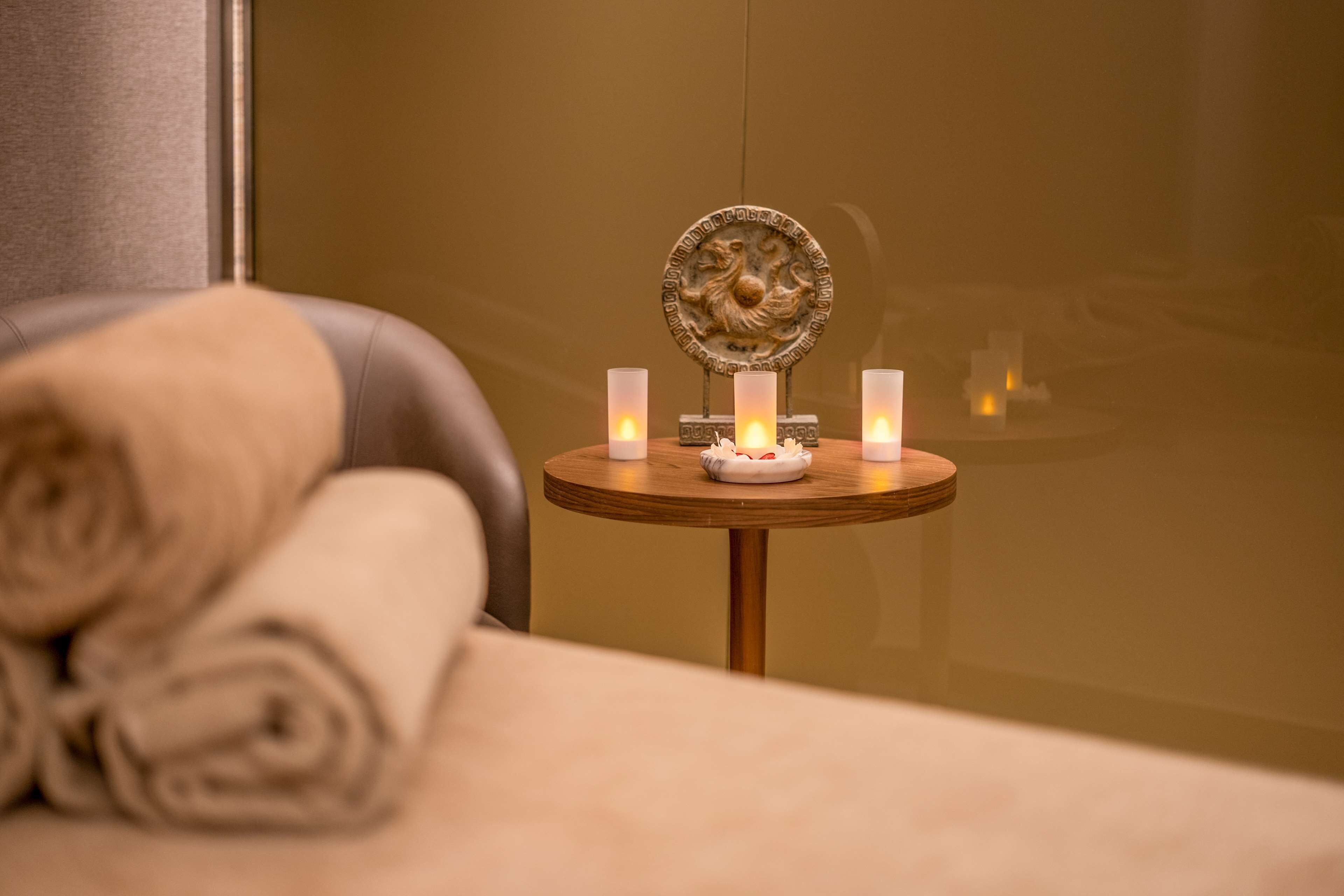 A neatly made bed with rolled towels, a small round table with candles, and a decorative plate.
