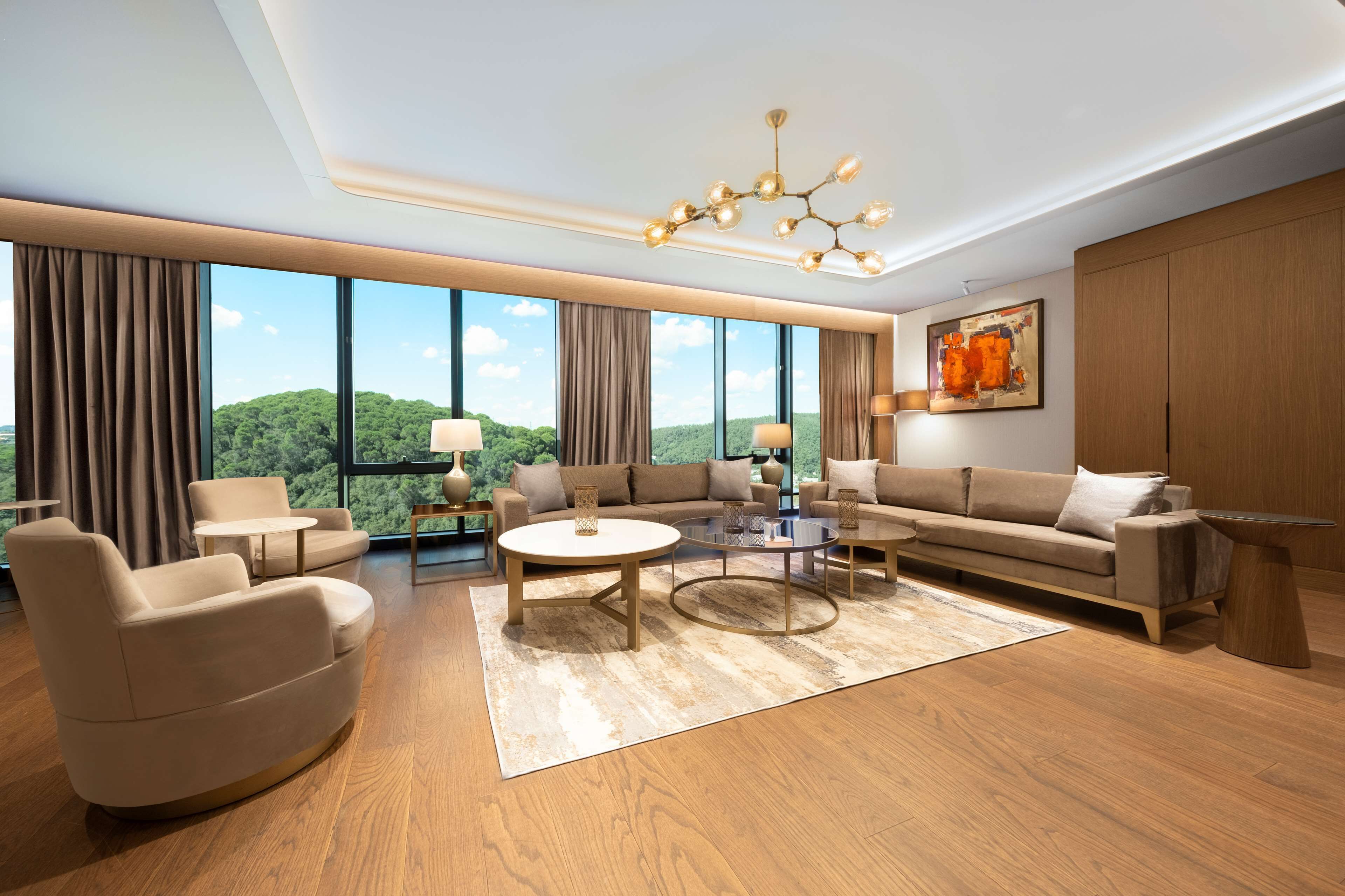 A spacious living room with a large window, a modern chandelier, and a view of a green landscape.