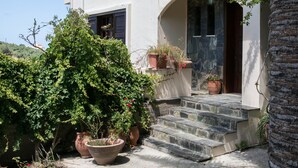 Una casa de dos pisos con una escalera de piedra que lleva a la entrada, rodeada de plantas en macetas y una palmera.
