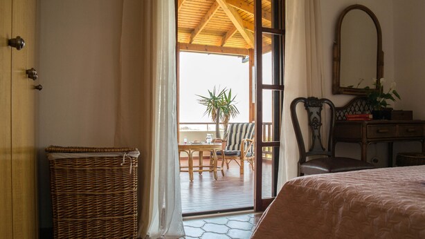 Un dormitorio con una cama, una cesta de mimbre, una silla de madera, una mesa con sillas y vistas al exterior.