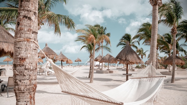 A beach scene with hammocks, palm trees, and thatched umbrellas.