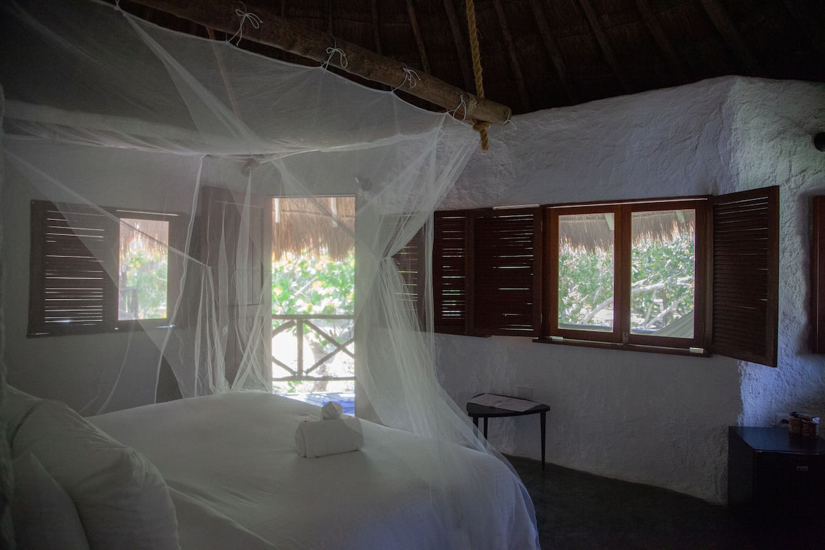 Une chambre avec un lit recouvert d’une moustiquaire, des volets en bois aux fenêtres et une petite table.