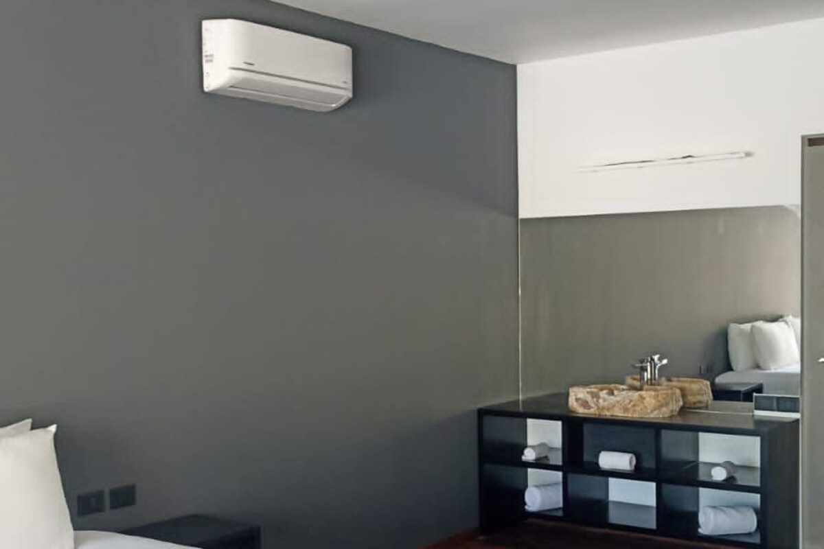 Una habitación de hotel moderna con una cama grande, suelo de madera oscura, pared gris, unidad de aire acondicionado blanca, cómoda de madera oscura con cajones y un espejo sobre la cómoda.