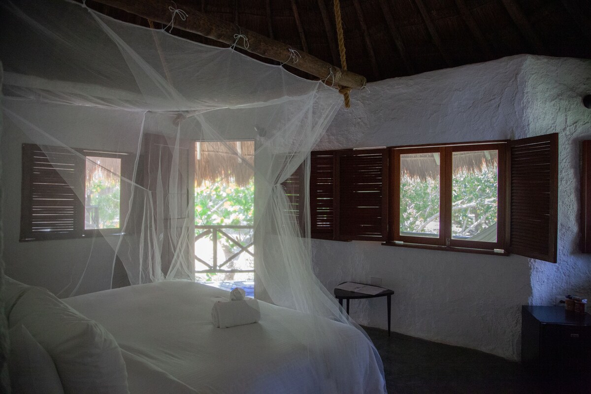 A room with a bed under a mosquito net, wooden shutters on windows, and a small table.