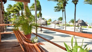 A wooden deck with a bench and potted plants, overlooking a beach with palm trees and thatched umbrellas.