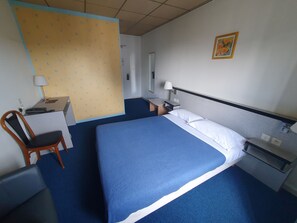 A hotel room with a bed, a desk, a chair, a nightstand, and a wall-mounted lamp. - Hôtel Terminus (Saint-Malo)