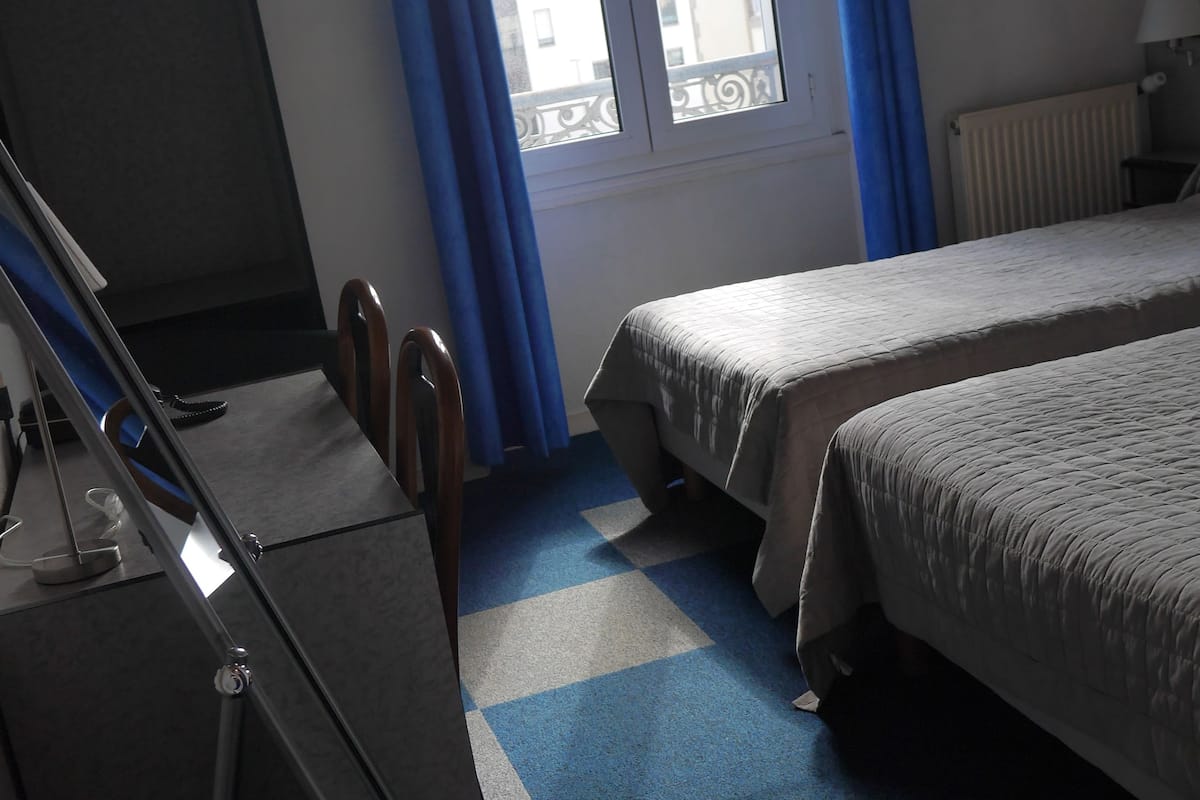 A hotel room with two beds, a desk, and a window with blue curtains.