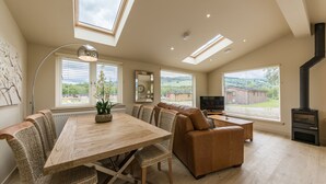 A spacious living room with a wooden dining table, wicker chairs, a brown leather sofa, a TV, and a wood-burning stove. - Balloch Park (Aberfeldy)