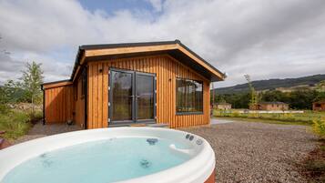 Una cabaña moderna de madera con hidromasaje en la fachada, rodeada por un sendero de grava y vegetación.