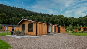 A wooden cabin with a gravel driveway, large windows, and a patio area with outdoor furniture. - Balloch Park (Aberfeldy)