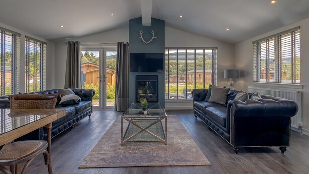A spacious living room with a fireplace, leather sofas, a glass coffee table, and large windows offering a view of the outdoors. - Balloch Park (Aberfeldy)