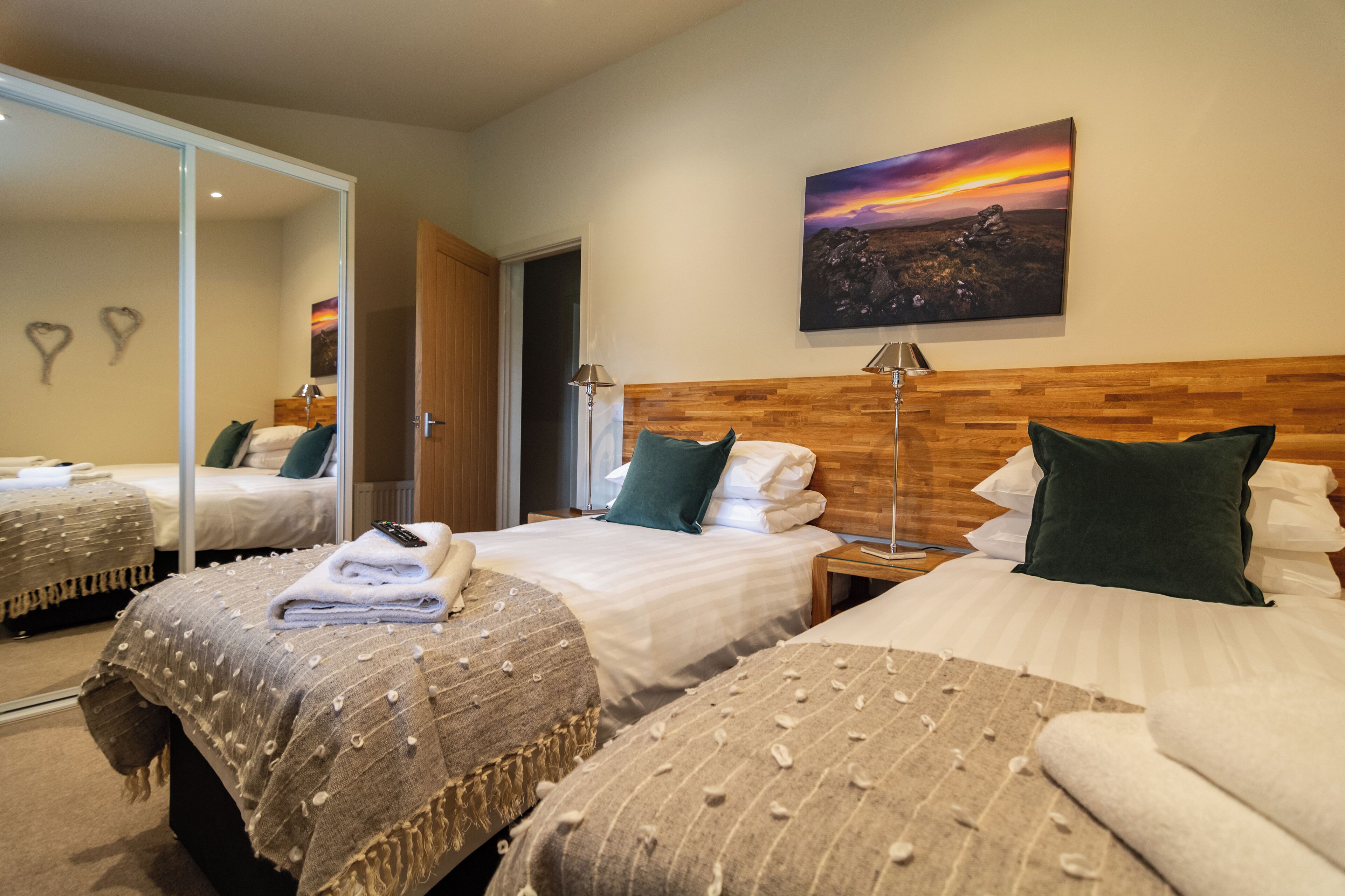 A hotel room with two beds, a wooden headboard, a painting of a sunset, and a mirror reflecting the room.