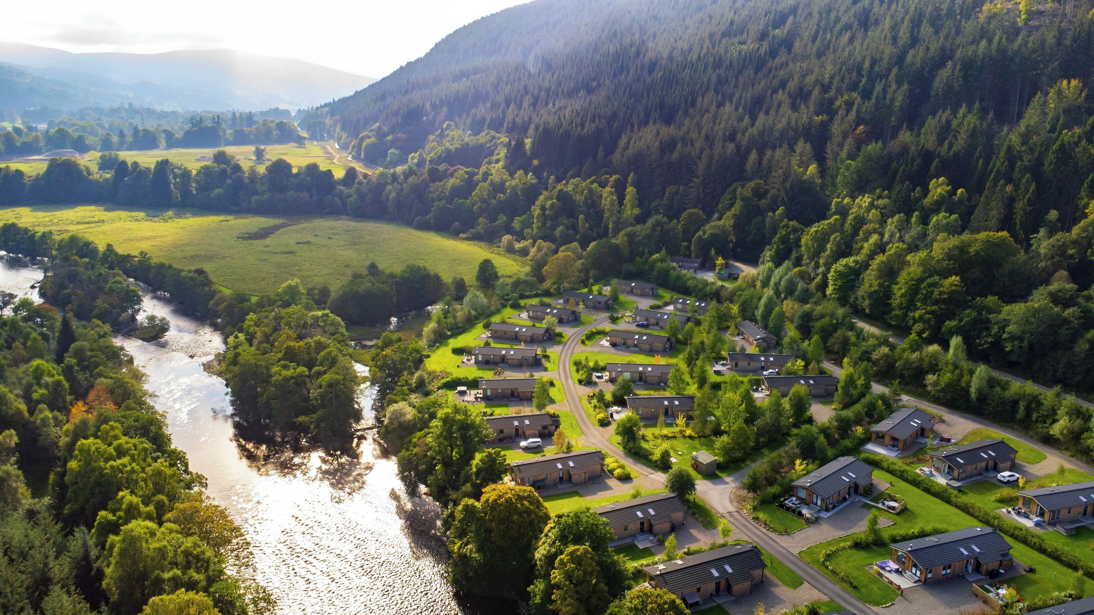Une vue aérienne d’un quartier résidentiel où les maisons sont disposées selon un plan en forme de grille, avec une rivière qui traverse la vallée et une colline boisée en arrière-plan.