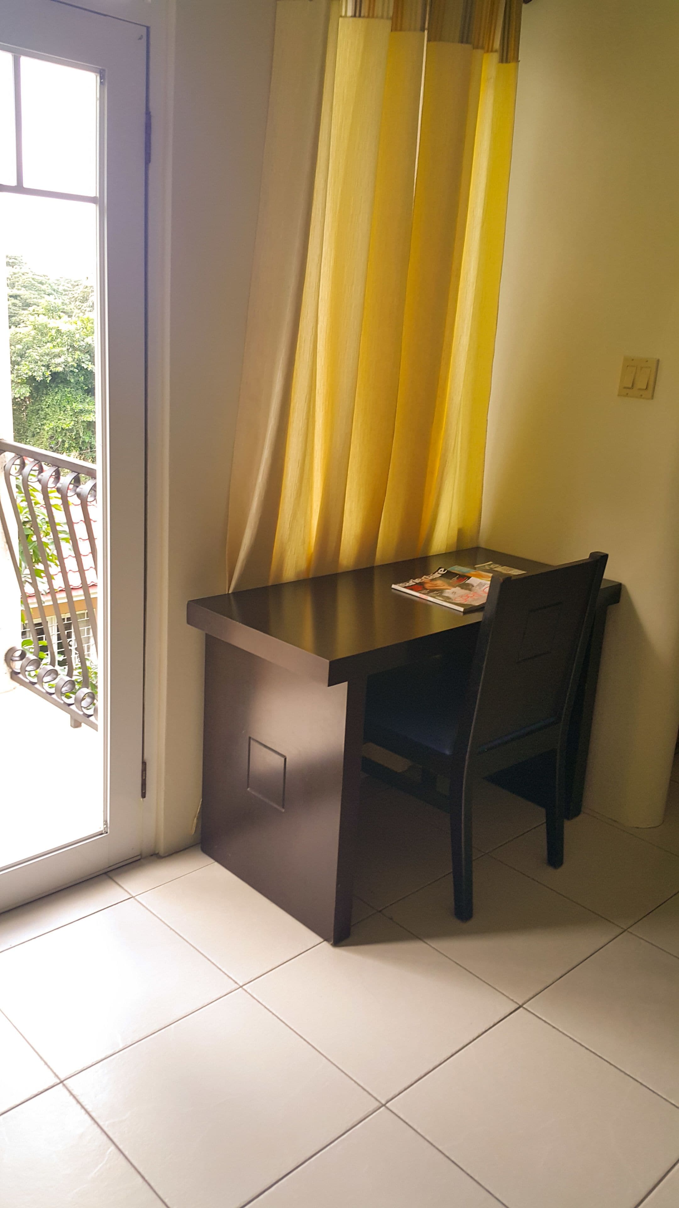 Une pièce avec un bureau et une chaise, une fenêtre avec des rideaux et un balcon avec une balustrade.