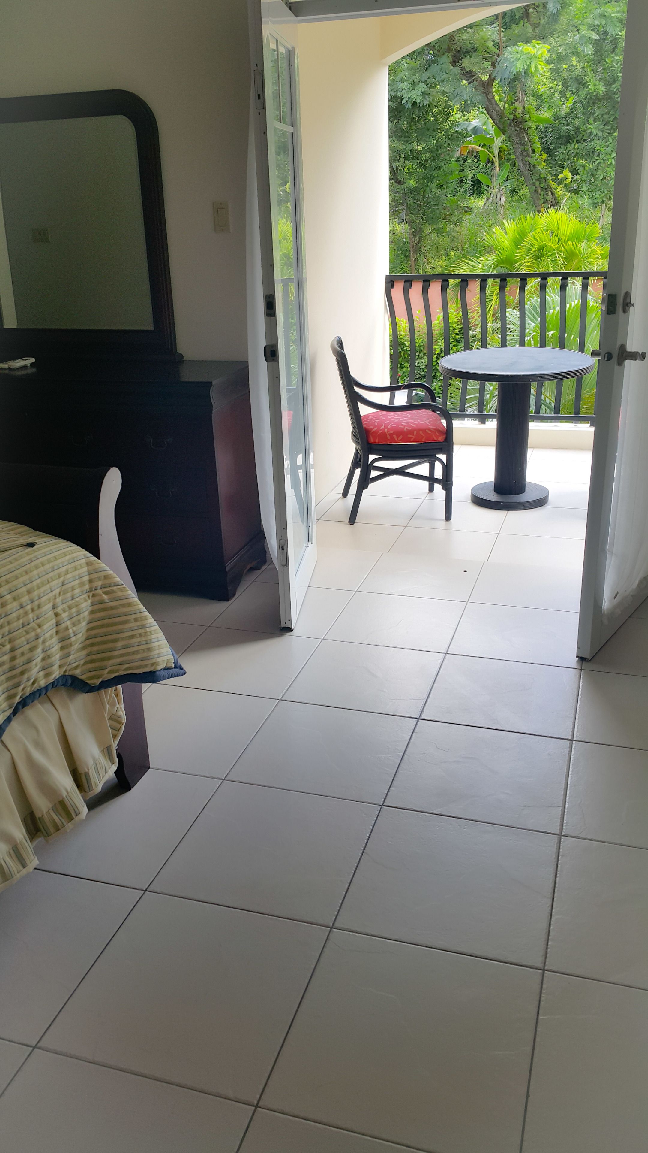 A hotel room with a bed, a TV, a balcony with a table and chair, and a view of greenery.