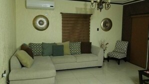 A living room with a beige sofa, patterned cushions, a patterned armchair, a wall-mounted air conditioner, and a chandelier. - The Hamptons of Acadia (Kingston)