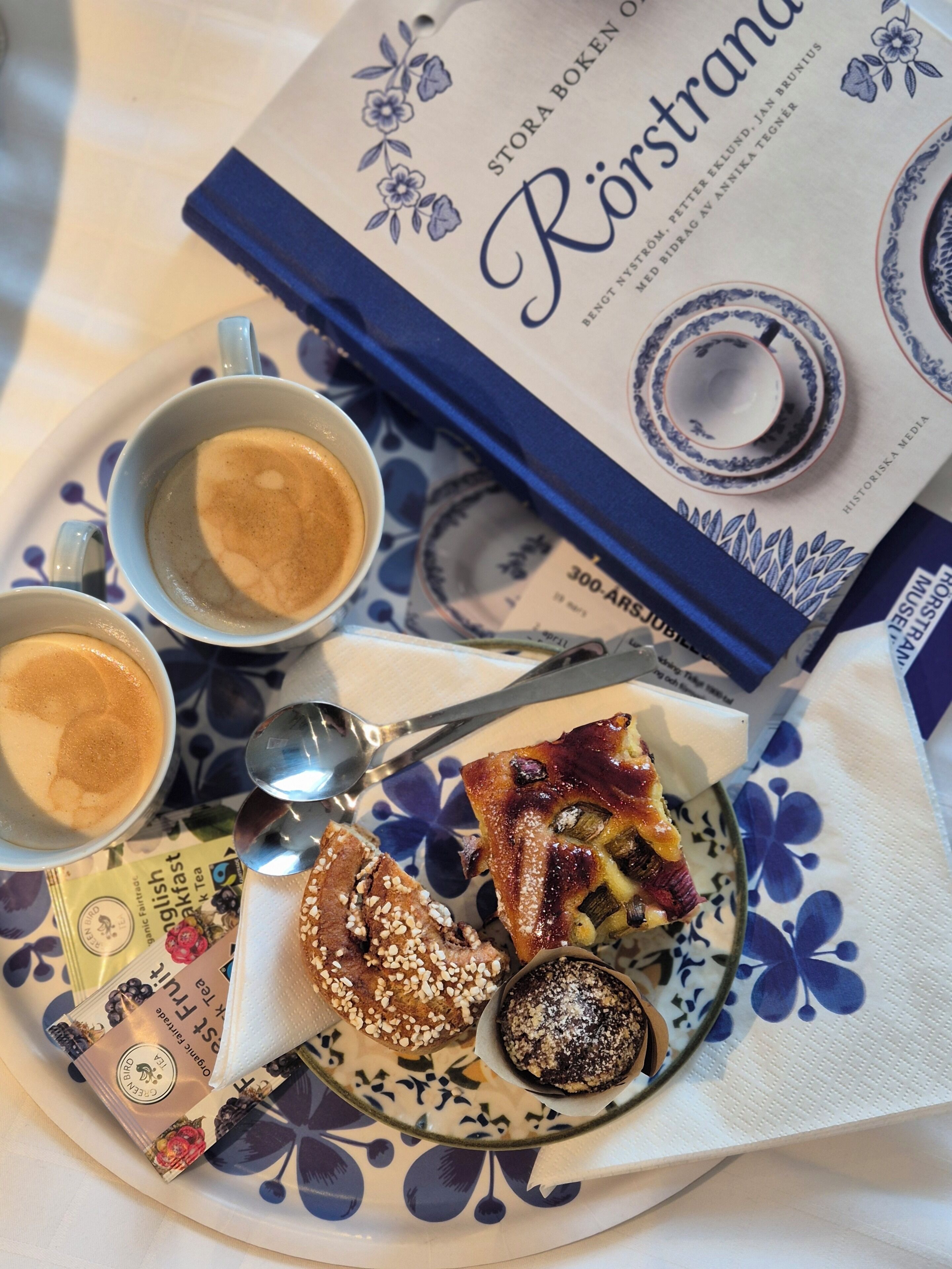 A tray with two cups of coffee, a plate with pastries, a spoon, and a book titled 'Rörstrand'.
