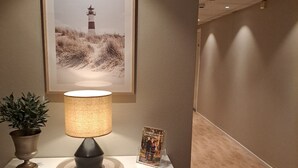 Una mesita de noche con una lámpara, un cuadro enmarcado de un faro, una planta pequeña en maceta y un atril con un libro.