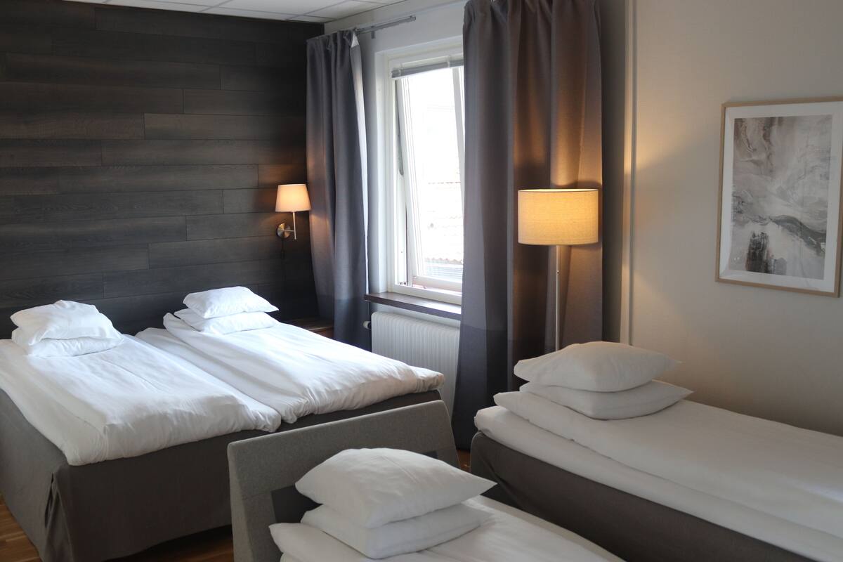 A hotel room with two beds, a window with curtains, a radiator, and two wall-mounted lamps.