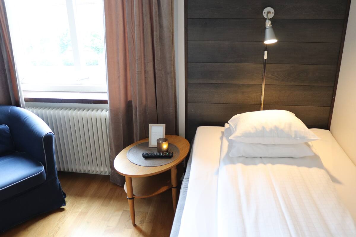 A hotel room with a bed, a small table with a remote control, a lamp, and a blue chair.
