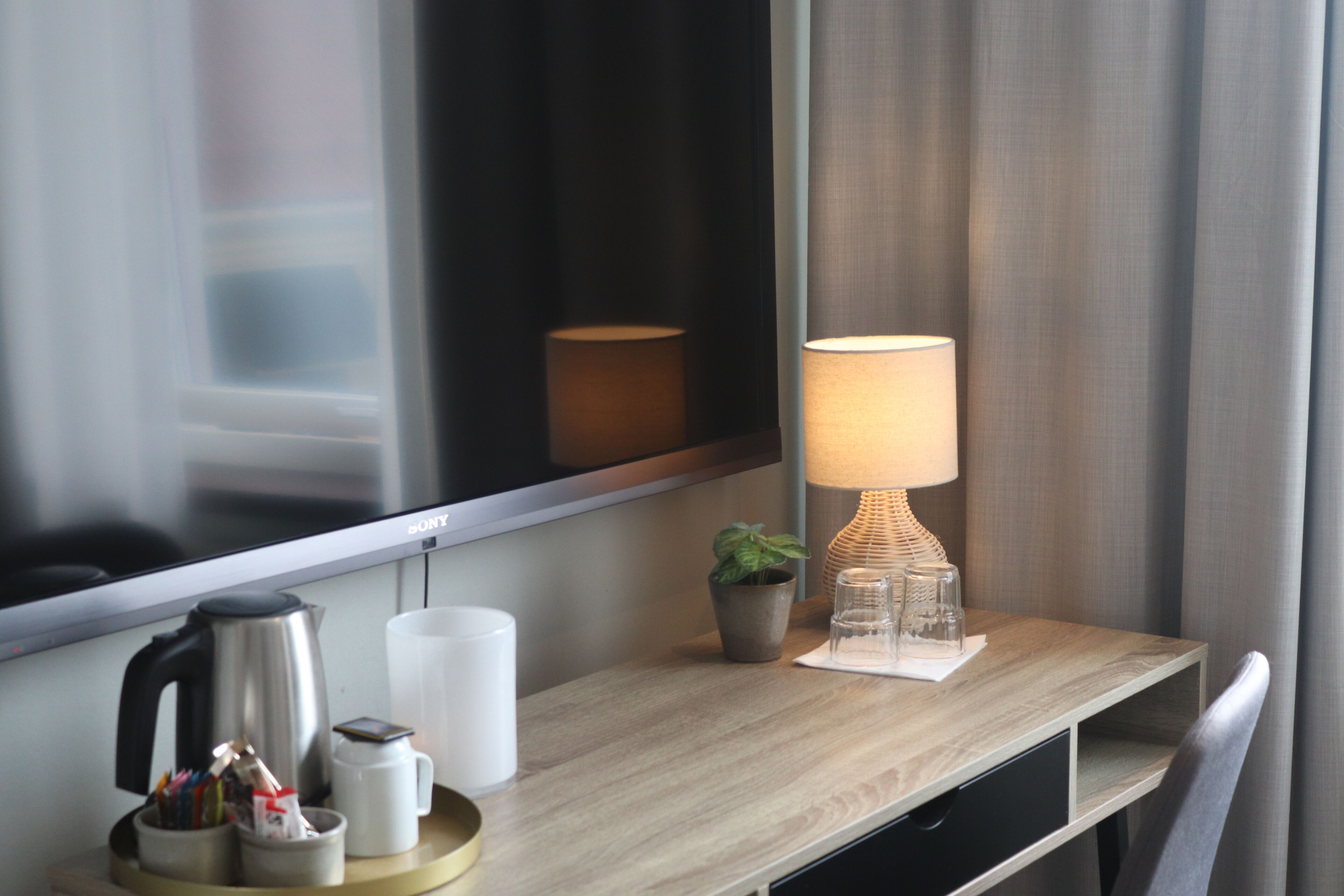Habitación de hotel con un escritorio de madera, una lámpara, una planta en maceta, una cafetera y un televisor.