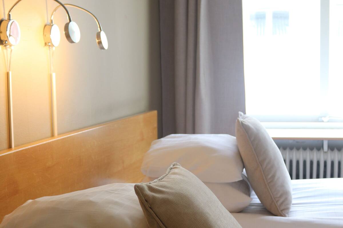 A neatly made bed with white and beige pillows, a wooden headboard, and a wall-mounted light fixture.