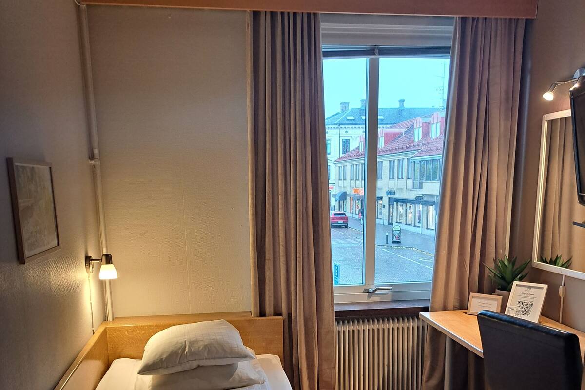 A hotel room with a bed, a desk, a chair, a radiator, and a window with curtains.