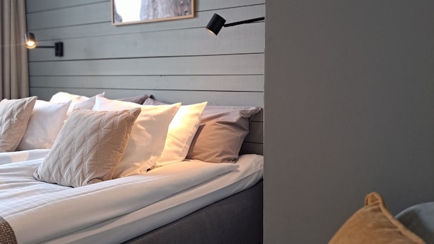 Una cama bien hecha con sábanas blancas y varios almohadones, una lámpara fijada a la pared y un cuadro enmarcado sobre la cama.