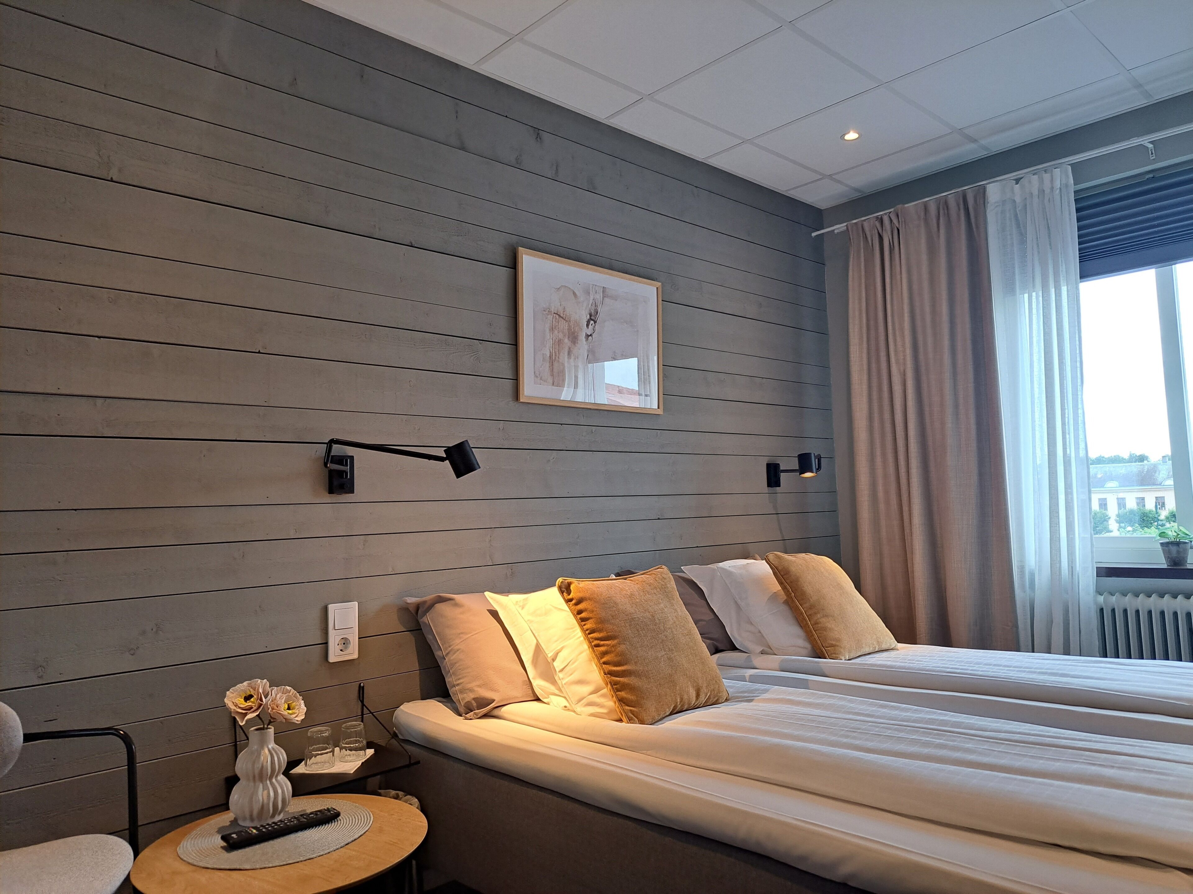 A hotel room with a large bed, a bedside table with a vase and glasses, a wall-mounted lamp, and a window with curtains.