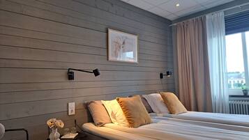 A hotel room with a large bed, a bedside table with a vase and glasses, a wall-mounted lamp, and a window with curtains.