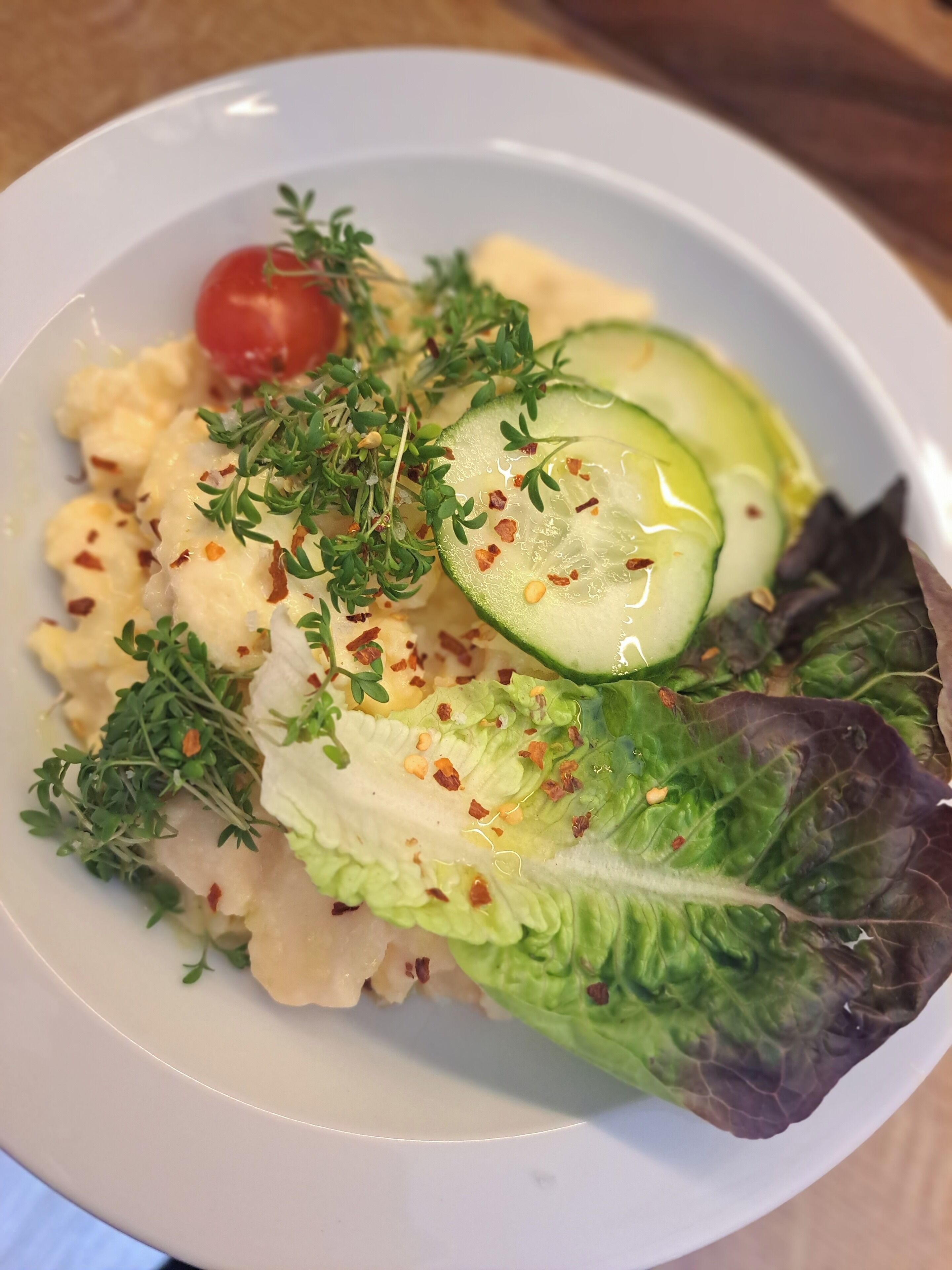 En vit tallrik med en sallad som innehåller salladsblad, skivor av gurka, en körsbärstomat och en garnering av örter.
