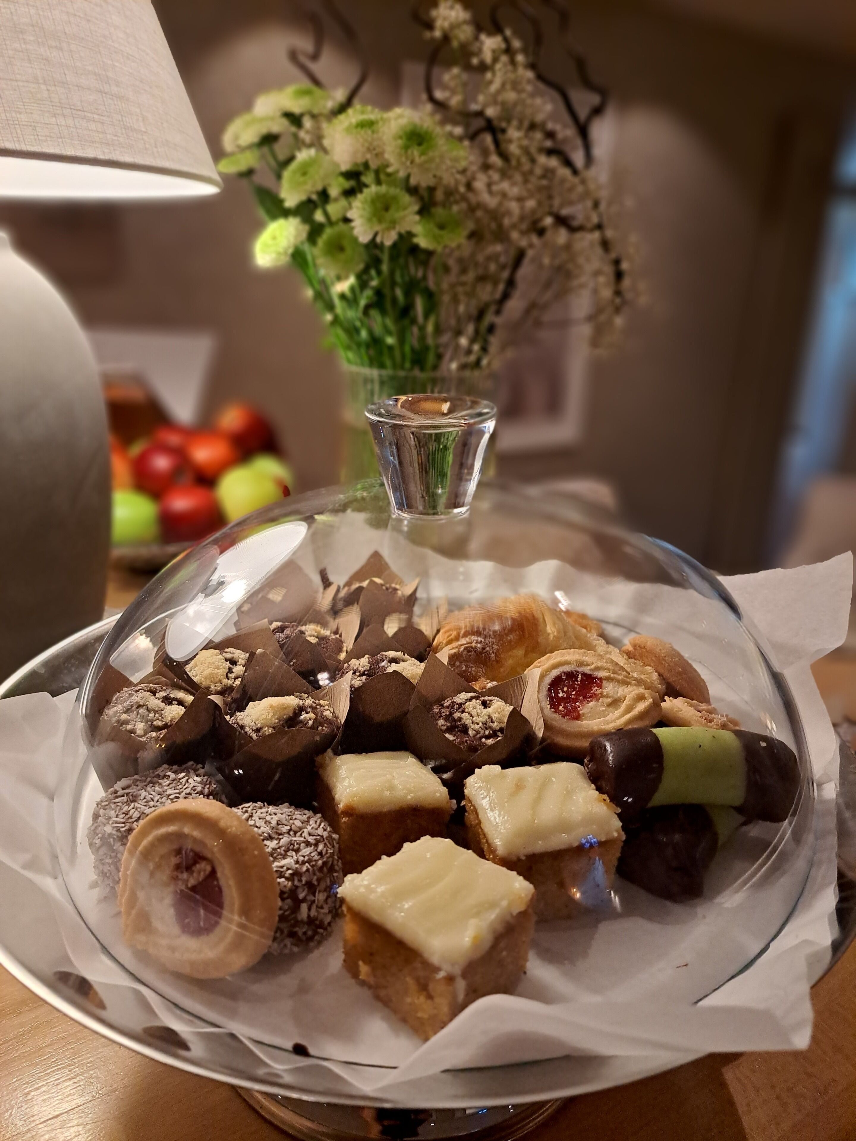 A dessert platter with various sweets on a glass plate, a vase with flowers, and a bowl of fruit in the background.
