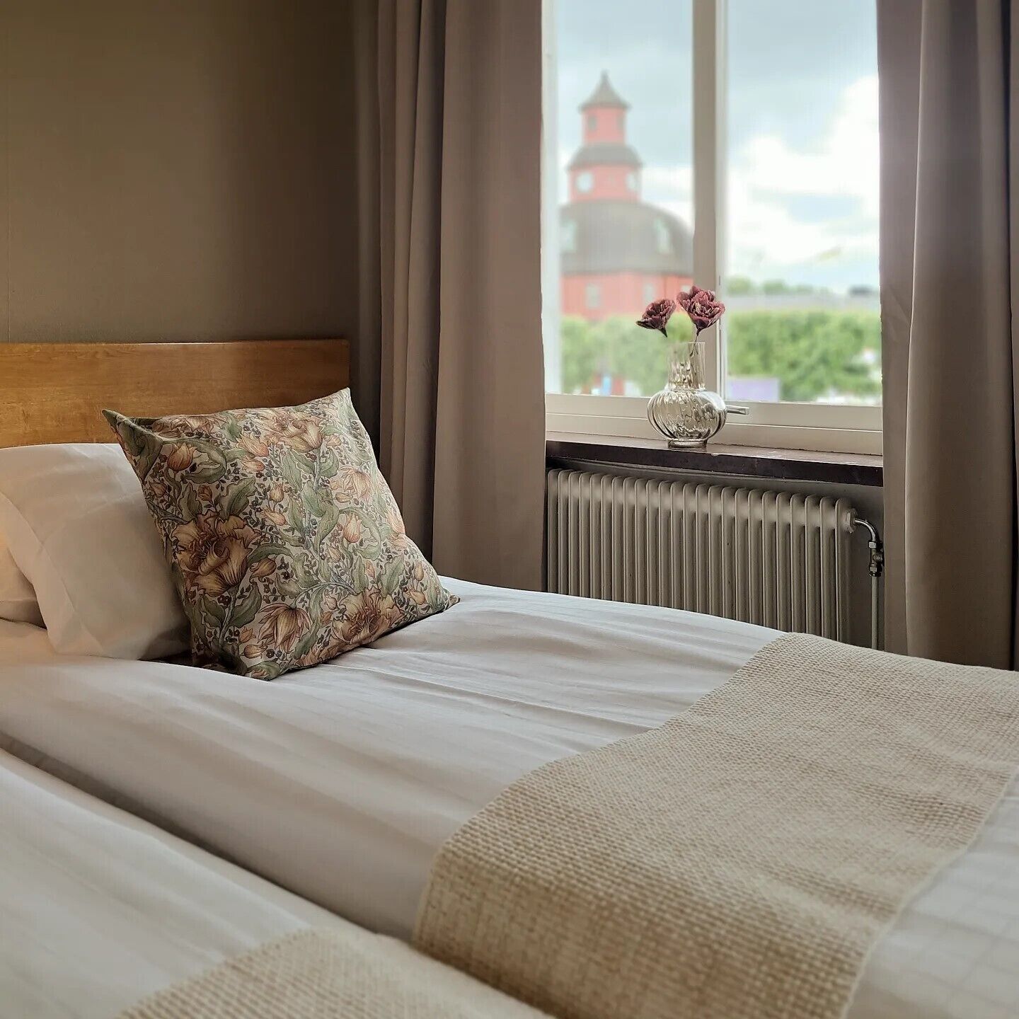 A neatly made bed with floral pillows, a vase with flowers, and a view of a lighthouse through a window.